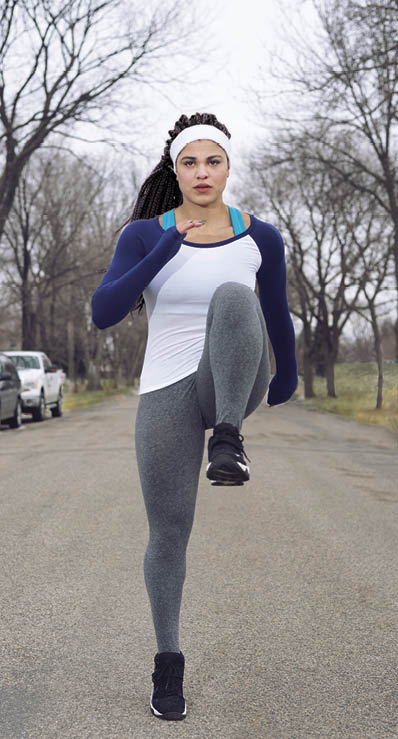 young woman warming up with knee raises knee ups in the middle of a suburban street   Facial expression is one of concentration, while dressed in sports wear 